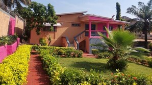 A two-story building with a pink facade, a covered staircase, and a well-maintained garden with various plants and a palm tree. - Villa Fatou Sow-Junior Suite-First Floor-R21 (Conakry)