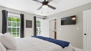 A bedroom with a bed, a ceiling fan, a TV, and a window with curtains.