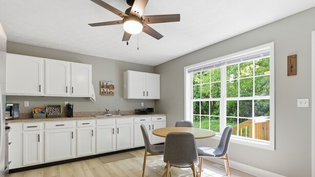 A modern kitchen with white cabinets, a dining table with chairs, a ceiling fan, and a large window with a view of greenery.