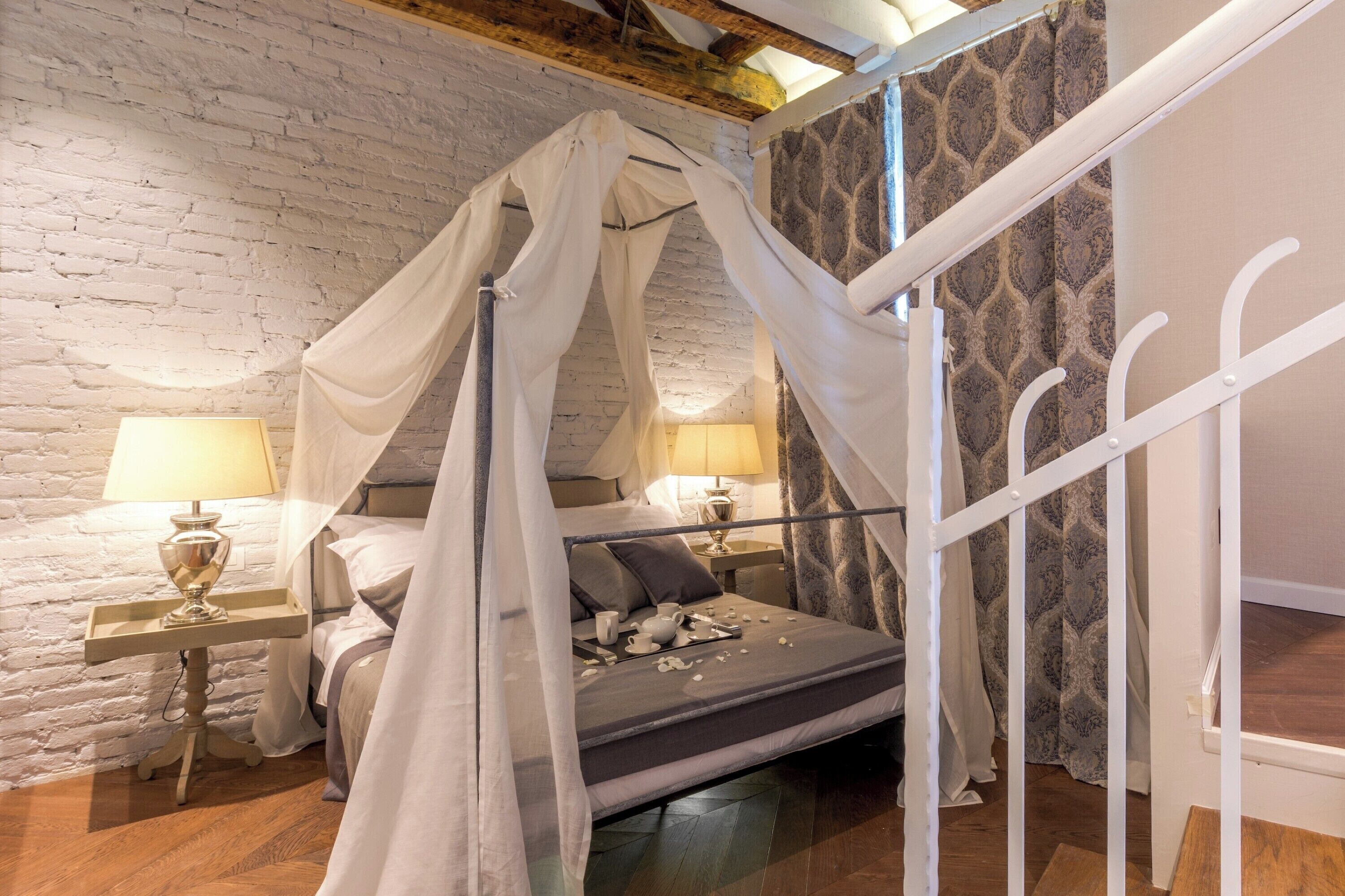 A four-poster bed with a canopy, two bedside lamps, a wooden floor, and a brick wall.