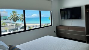 A hotel room with a large window offering a view of the ocean and palm trees, a bed, a TV mounted on the wall, and a wooden cabinet. - Villa Paradiso (San Juan)