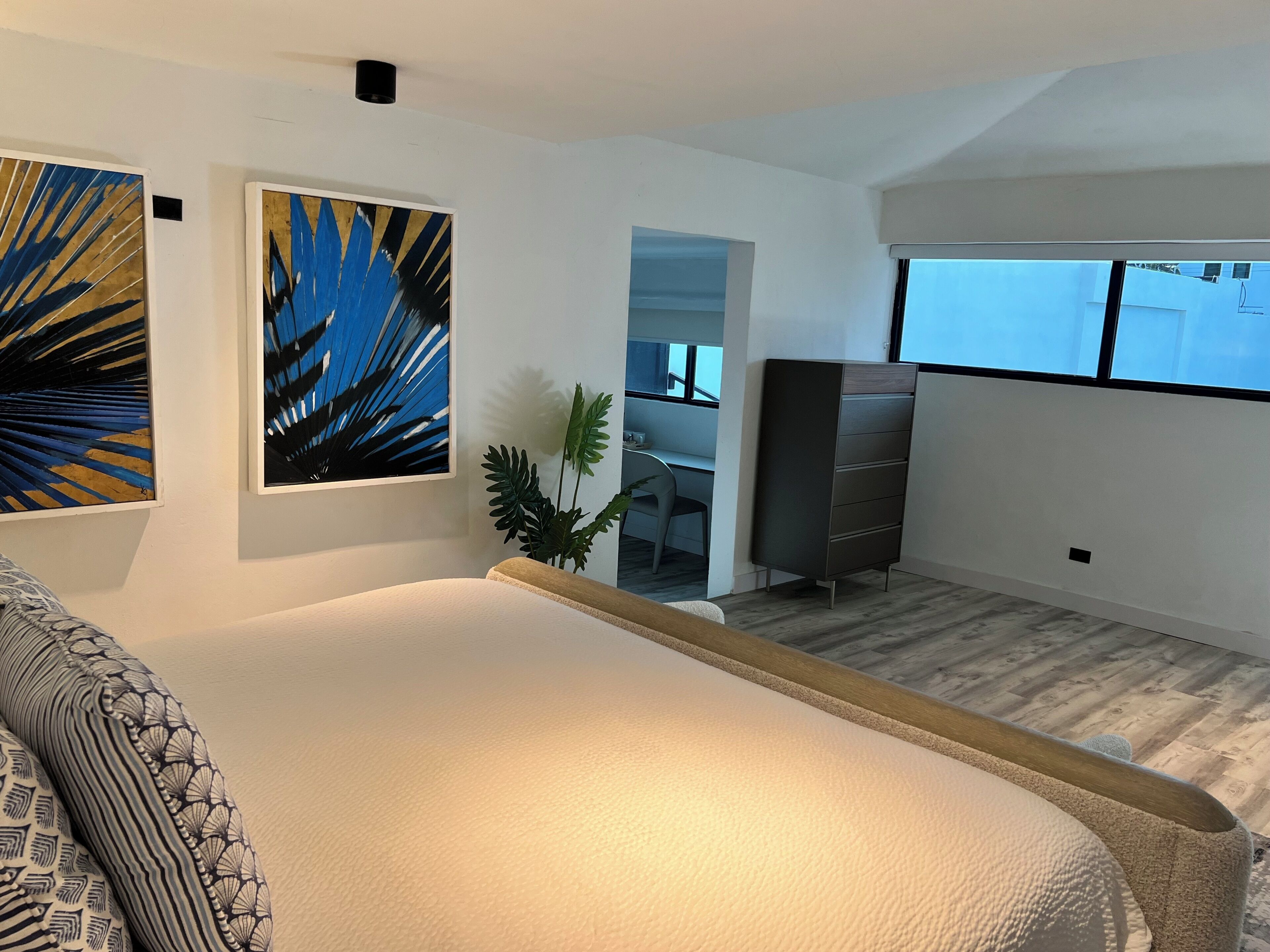 Une chambre à coucher comprenant un lit, un bureau, une chaise, une commode et deux tableaux abstraits.