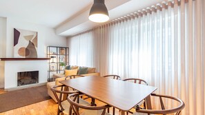 A modern living room with a wooden dining table, chairs, a sofa, a fireplace, and a large window with sheer curtains. - Stylish 3BR in Central Porto w/ AC by LovelyStay (Porto)