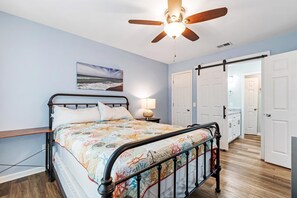 Ein Schlafzimmer mit Bett, Deckenventilator, Nachttisch, Lampe, einer Tür und einem Flur.