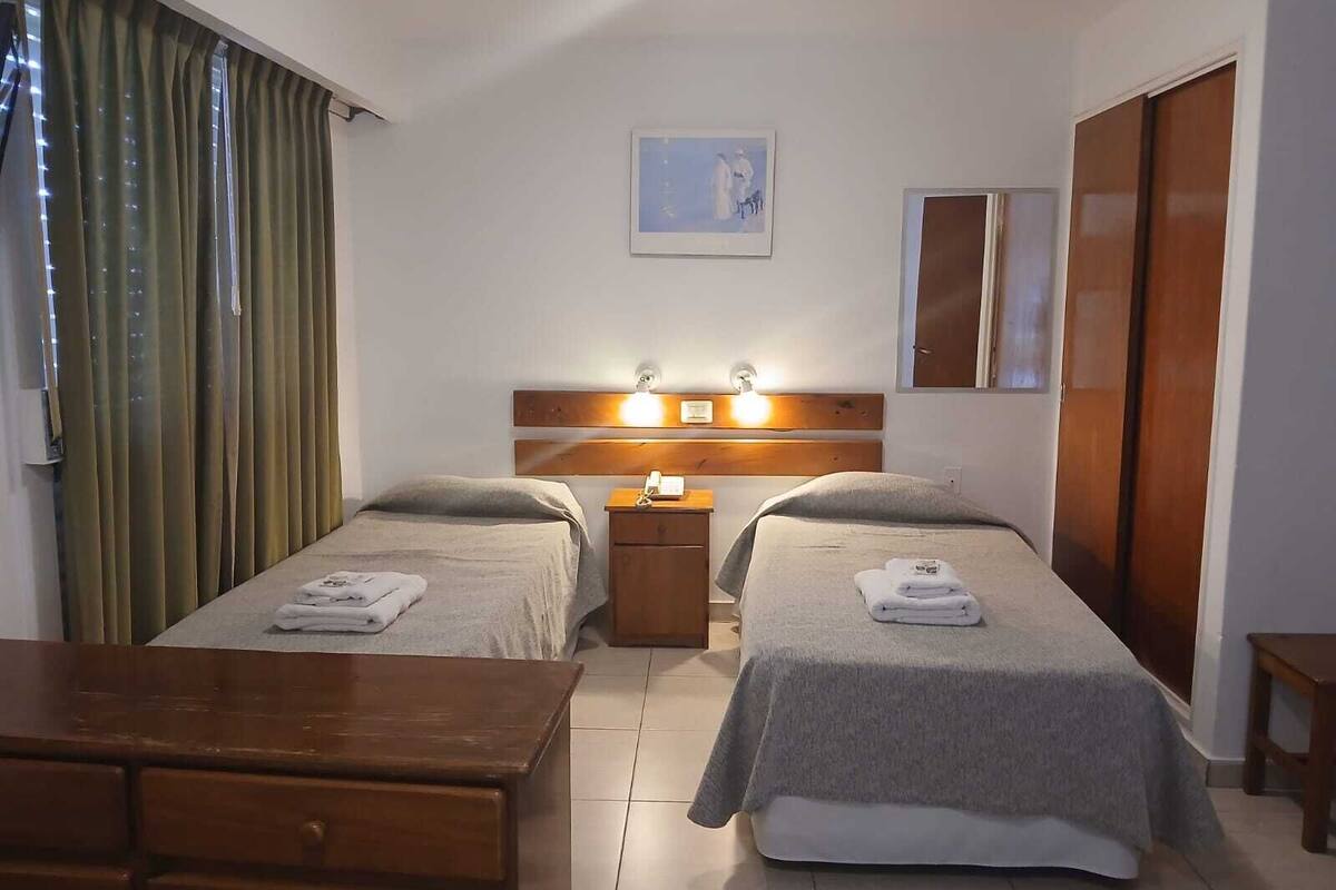Habitación de hotel con dos camas, un aparador de madera, una mesita de noche con lámpara y un cuadro en la pared.