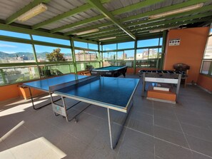 Ein Billardtisch, ein Kicker und ein Snookertisch in einem Raum mit großen Fenstern und Blick auf Gebäude und Berge.
