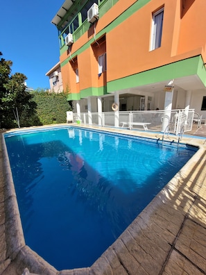 A swimming pool with a wooden deck, surrounded by a building with balconies and air conditioning units. - El Mirador (Villa Carlos Paz)
