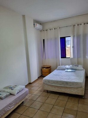 A bedroom with two beds, a window with curtains, a wooden nightstand, and an air conditioning unit. - Barra Lighthouse. SPACIOUS. Feet in the sand. SEA VIEW. (Salvador)
