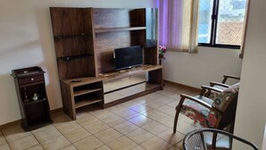Una sala de estar con un televisor sobre un mueble de madera, un archivador pequeño, una silla y un ventanal con cortinas.