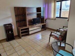 A living room with a TV on a wooden stand, a small cabinet, a chair, and a window with curtains. - Barra Lighthouse. SPACIOUS. Feet in the sand. SEA VIEW. (Salvador)