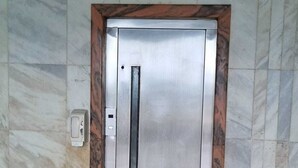Puerta de ascensor metálica con un tirador vertical, ubicada en una pared con azulejos de diseño similar al mármol.