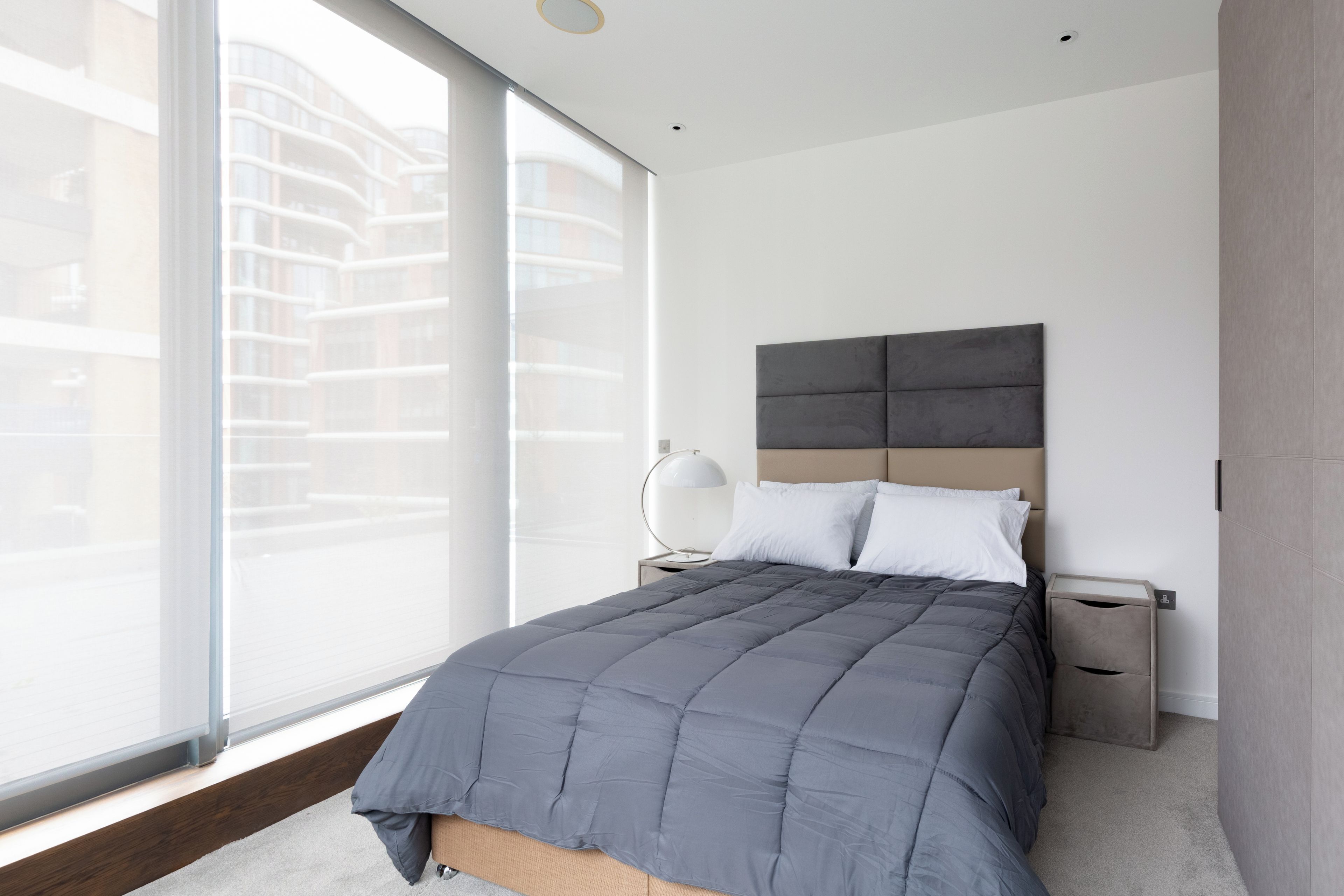 Una camera da letto moderna con un'ampia finestra, un letto con copriletto grigio, due comodini e una lampada a parete.