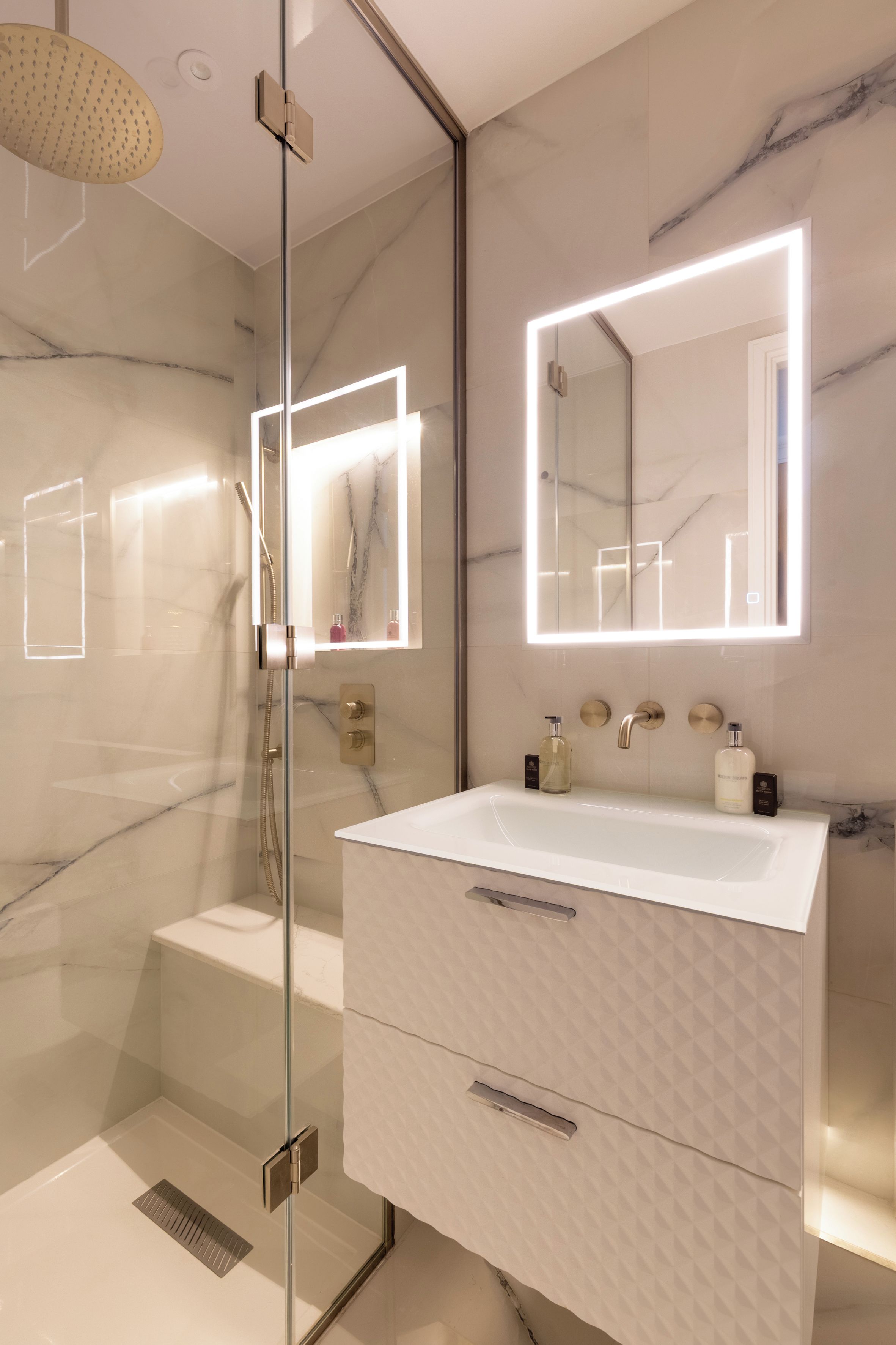 Un bagno moderno con cabina doccia in vetro, un lavabo bianco con piano in marmo e un grande specchio con illuminazione integrata.