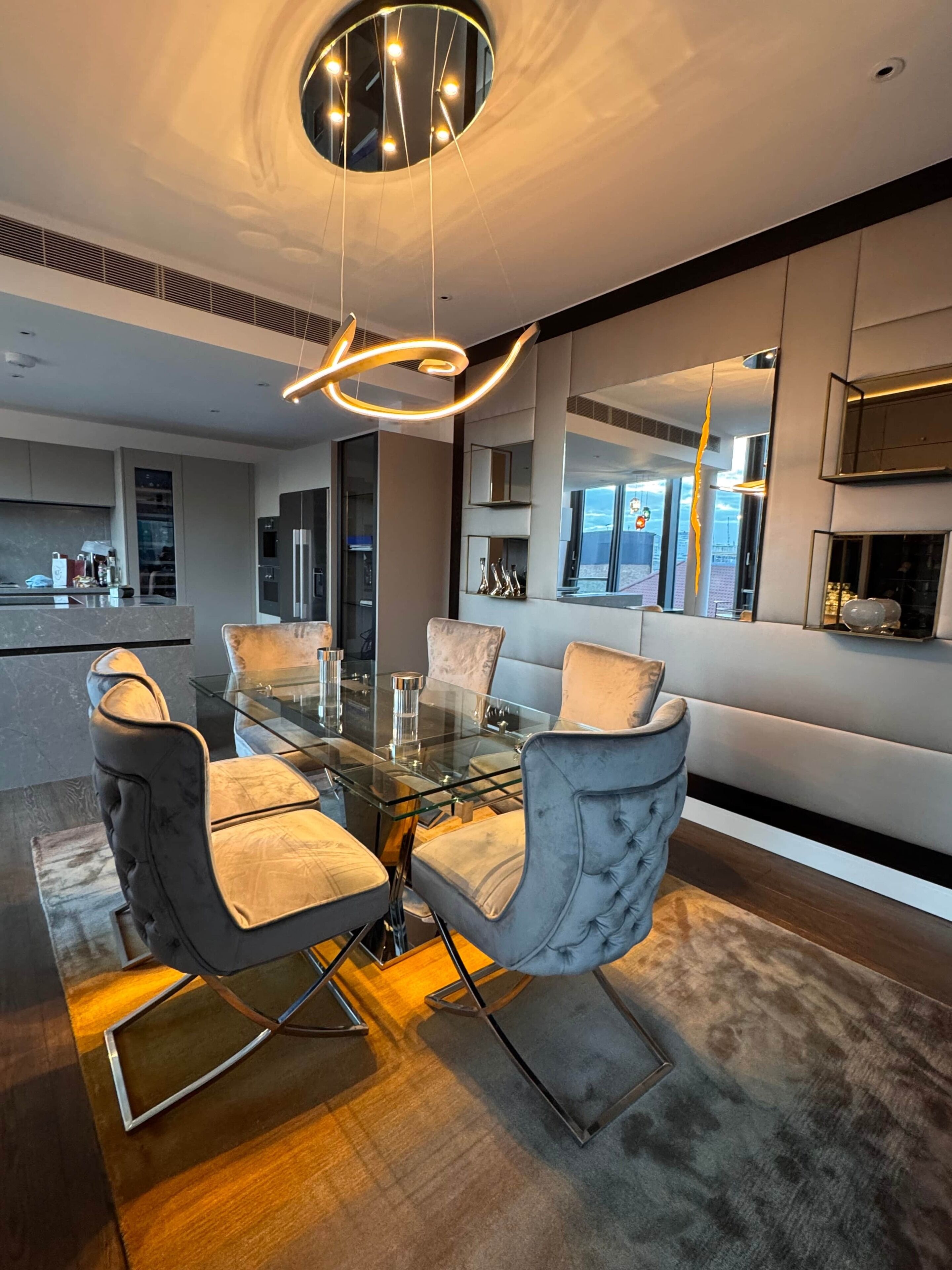 A modern dining area with a glass table and cushioned chairs, a contemporary light fixture, and a view of the city.