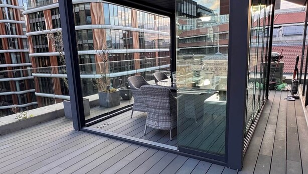 En moderne balkon med glasfacade, fletmøbler og udsigt til omkringliggende bygninger.
