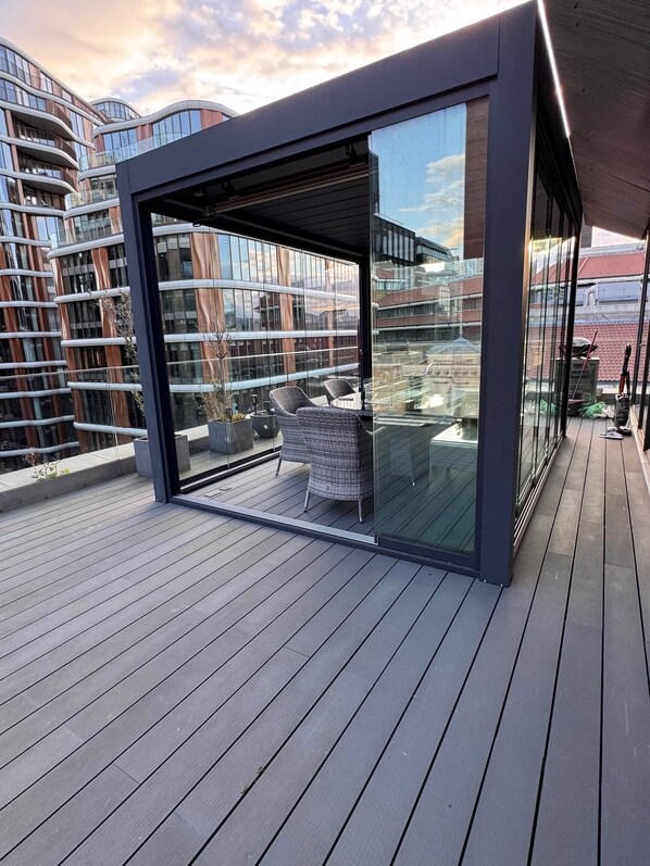A modern outdoor balcony with a glass enclosure, wicker furniture, and a view of surrounding buildings. - Luminar Aurelia suite,
Luxury Penthouse (London)