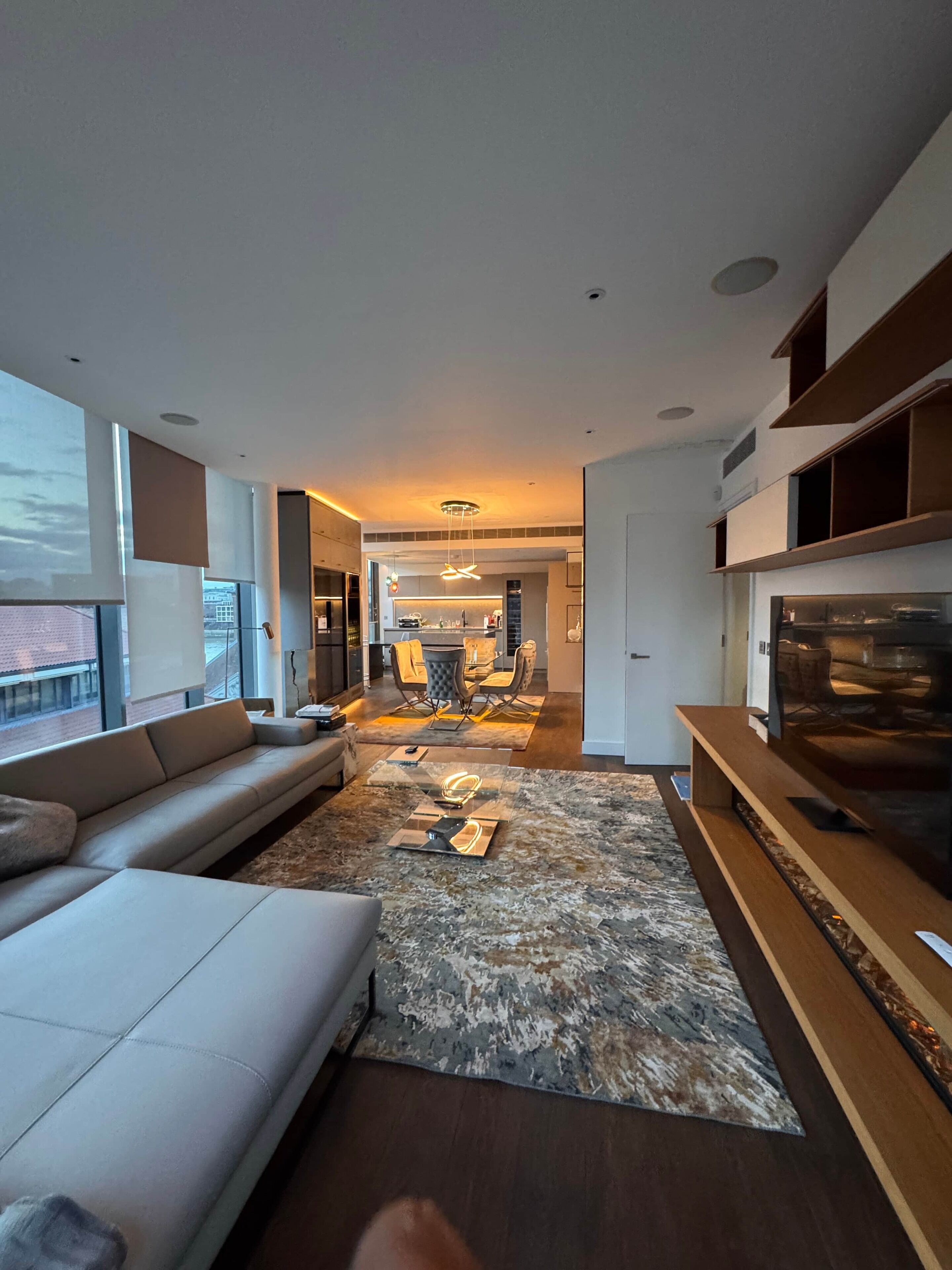 A modern living room with a sofa, a coffee table, and a TV stand. There is a dining area in the background with a table and chairs.