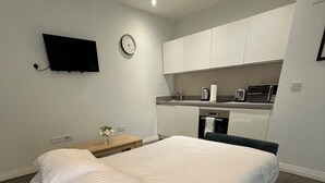 Une chambre moderne avec un grand lit, une petite table de chevet, un téléviseur fixé au mur et une kitchenette équipée d’un four à micro-ondes et d’une bouilloire.