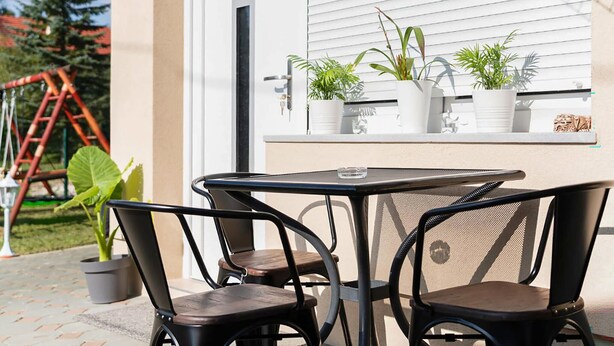 Eine Terrasse im Freien mit Tisch und Stühlen, Blumentöpfen und einem Fenster mit Jalousien.
