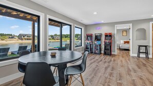 Espaço de refeições com grande mesa e cadeiras, jogos de fliperama e vista para um lago.