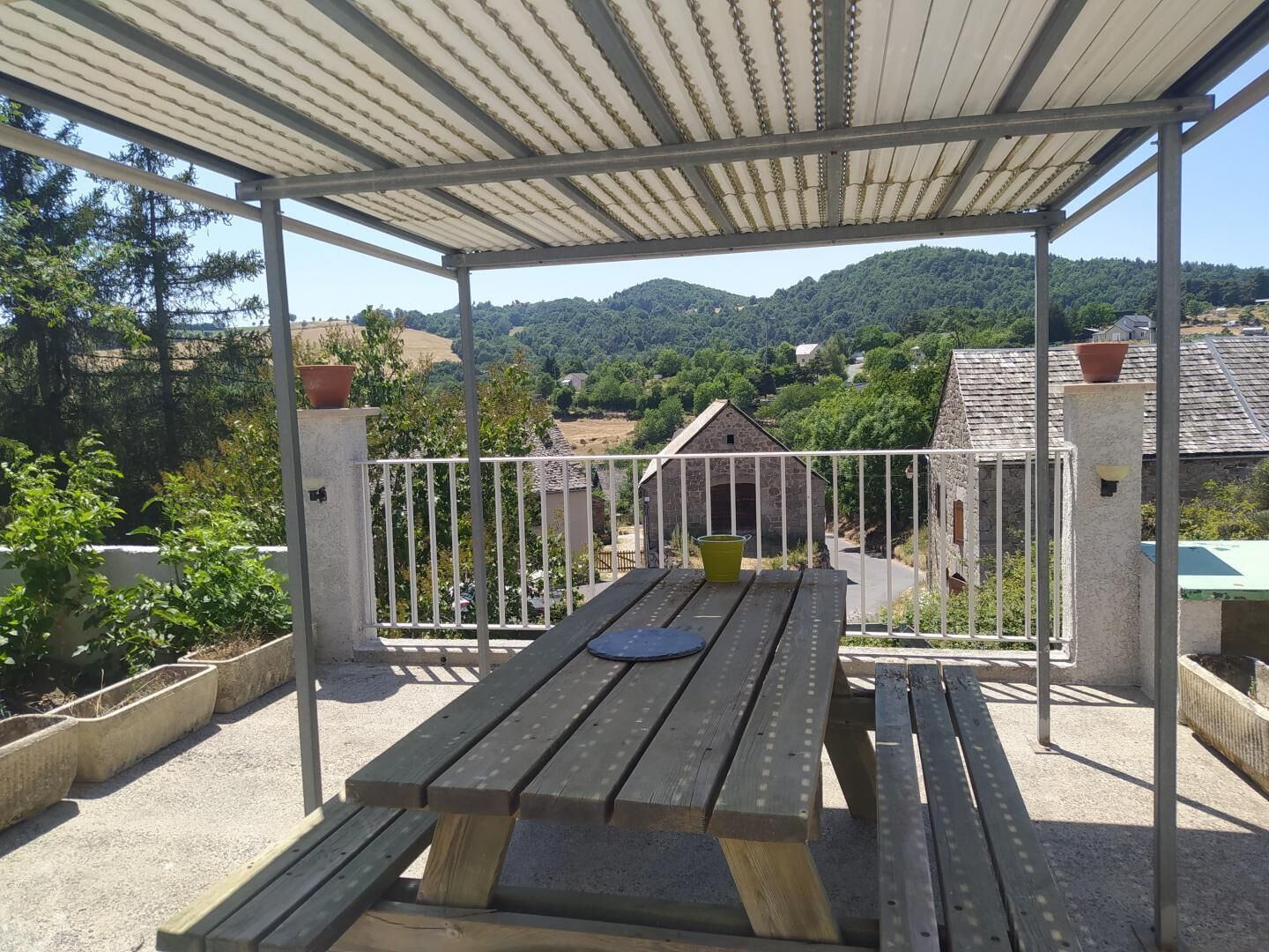 A covered outdoor area with a picnic table, a metal railing, and a view of a rural landscape with houses and hills.