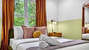 Ein ordentlich bezogenes Bett mit Kissen und einem zusammengerollten Handtuch, ein Nachttisch mit Lampe sowie ein Fenster mit Vorhängen.