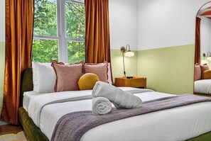 Ein ordentlich bezogenes Bett mit Kissen und einem zusammengerollten Handtuch, ein Nachttisch mit Lampe sowie ein Fenster mit Vorhängen.