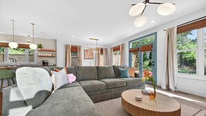 Ein modernes Wohnzimmer mit einer Eckcouch, einem runden Couchtisch, Pendelleuchten und großen Fenstern.