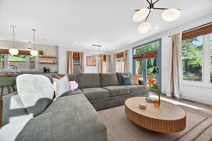 Ein modernes Wohnzimmer mit einer Eckcouch, einem runden Couchtisch, Pendelleuchten und großen Fenstern.