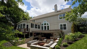 Una casa de dos pisos con un patio que cuenta con mobiliario exterior, un brasero y una chimenea de piedra.