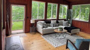 Una habitación acogedora con techo de madera, un sofá con cojines, una mesa de centro y grandes ventanales que ofrecen vistas al verdor.