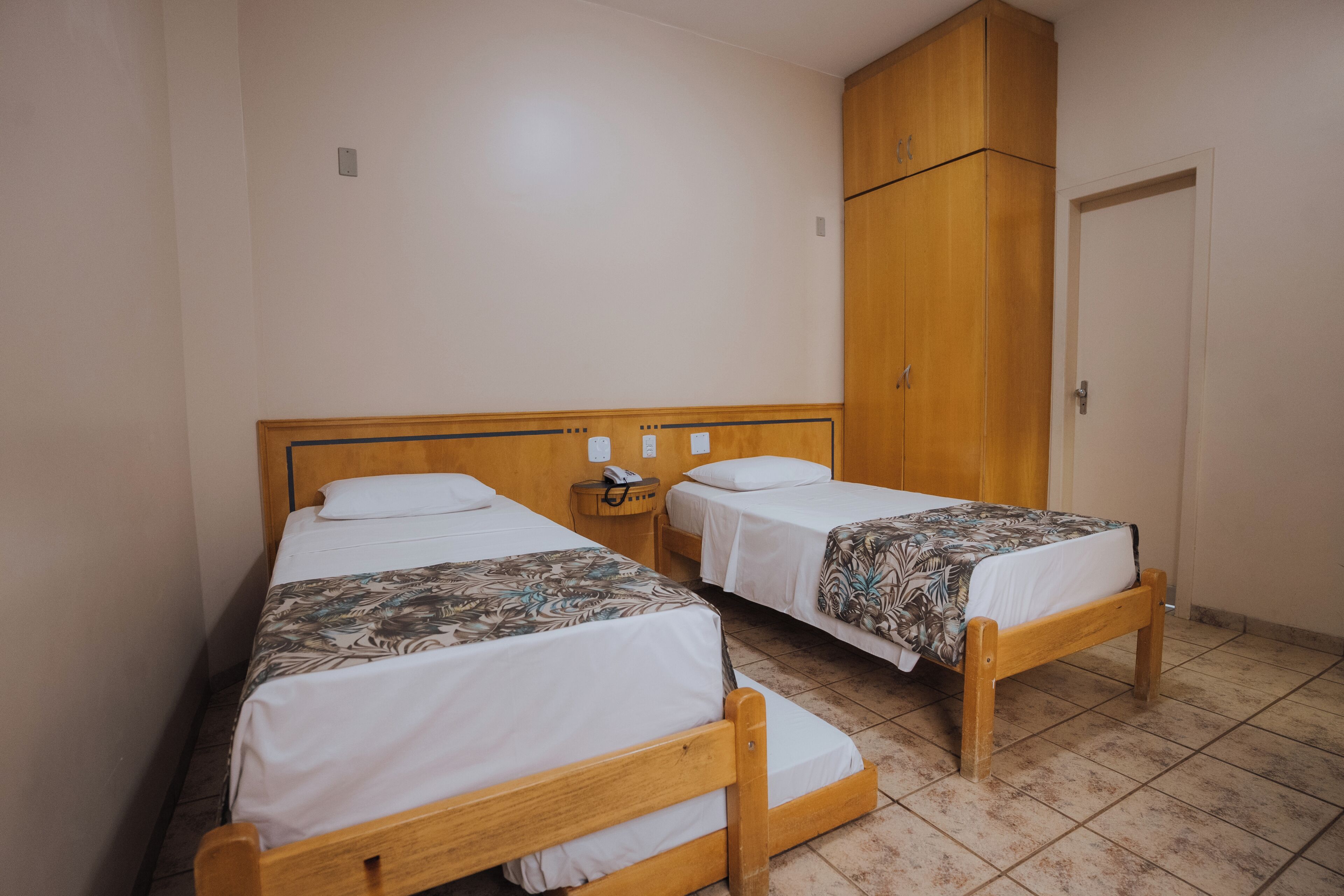 Two single beds with wooden frames, a wooden wardrobe, and a tiled floor.
