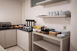 A kitchenette with a coffee maker, tea kettle, and a fruit bowl. There are shelves with plates and cups, and a basket with utensils. - Hotel Nacional Service (Goiania)
