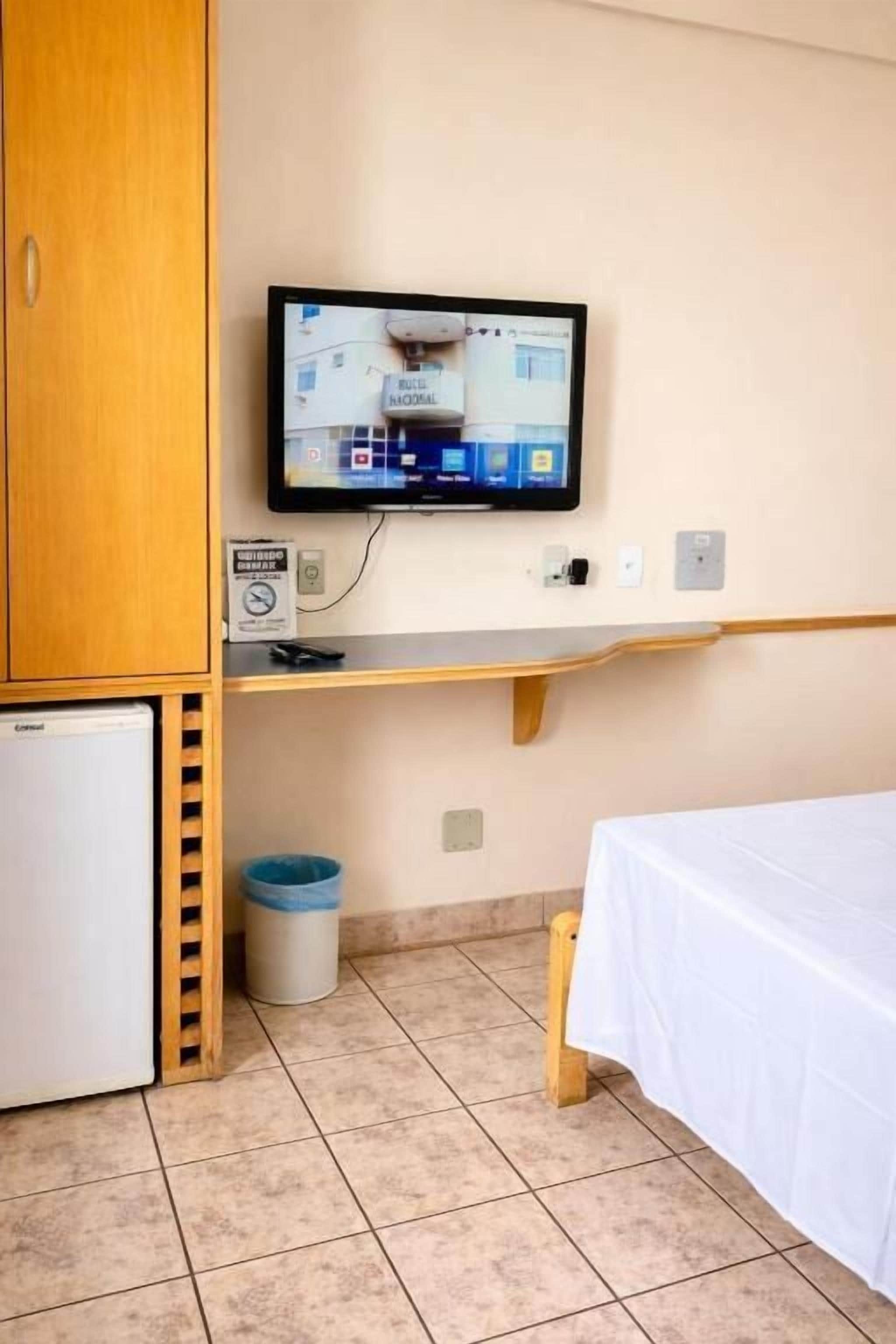 Una habitación con una cama, un televisor montado en la pared, un refrigerador pequeño, un cubo de basura y un armario de madera.