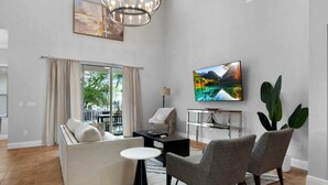 Ein modernes Wohnzimmer mit einer Couch, Sesseln, einem Couchtisch, einem Fernseher und einem großen Fenster mit Vorhängen.