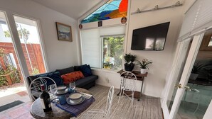 Una sala de estar acogedora con un sofá, una mesa de comedor con sillas, un televisor montado en la pared y una ventana con persianas.