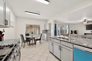 A modern kitchen with a gas stove, white cabinets, a dining area with chairs, and a view to the outside. - Large 6BR 3BA Home Near AT&T Stadium & Six Flags (Grand Prairie)