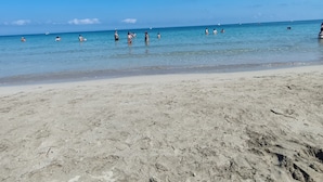 Une plage avec une eau d’un bleu limpide, un rivage sablonneux et quelques personnes qui se baignent.