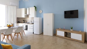 Une cuisine moderne avec des armoires blanches, un four encastré et un four à micro-ondes. On y trouve un coin repas avec une table et des chaises, ainsi qu’un téléviseur fixé au mur.