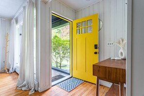 Une pièce avec un plancher en bois, une porte jaune, une table d’appoint en bois et un vase blanc contenant des plantes séchées.