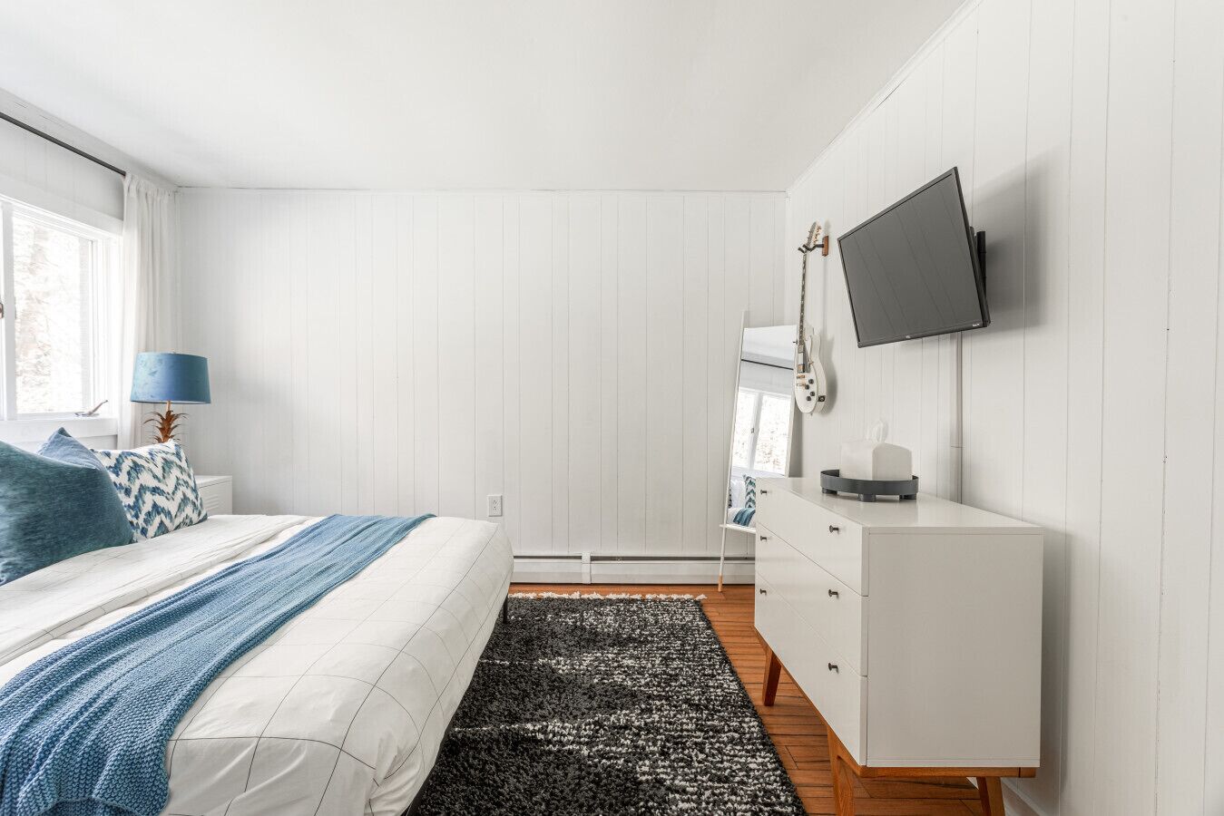 Quarto com cama, televisão fixada na parede, penteadeira e janela com cortinas.
