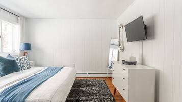 Quarto com cama, televisão fixada na parede, penteadeira e janela com cortinas.