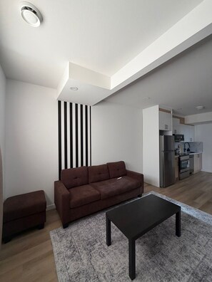 Ein modernes Wohnzimmer mit Sofa, Hocker und Couchtisch, im Hintergrund eine Küche und eine gestreifte Wand.