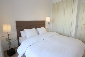 Een net opgemaakt bed met witte lakens, een glazen bijzettafeltje met een lamp en een kledingkast met een lichtgekleurde deur.