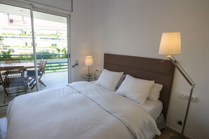 A hotel room with a bed, a table with chairs, a lamp, and a view of greenery outside. - Apartment Parc Del Mar (Sitges)