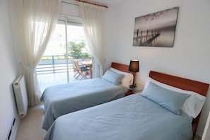 A hotel room with two beds, a table with chairs, a lamp, and a view of the outdoors. - Apartment Parc Del Mar (Sitges)