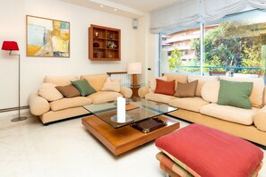 Una sala de estar con dos sofás, una mesa de centro de vidrio, una lámpara de pie roja, una estantería y un ventanal con vistas a zonas verdes.