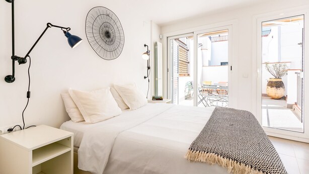 A bedroom with a bed, bedside table, lamp, wall art, and a view of the outside. - Casa Cipres - Three Bedroom Apartment, Sleeps 6 (Sitges)