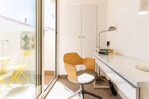 Habitació moderna amb una porta de vidre que dona a un balcó, una cadira de fusta, un escriptori amb una làmpada i una petita biblioteca.