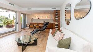 Un salon moderne avec une table basse en verre, un fauteuil en cuir, un canapé blanc agrémenté de coussins et, au fond, une cuisine.